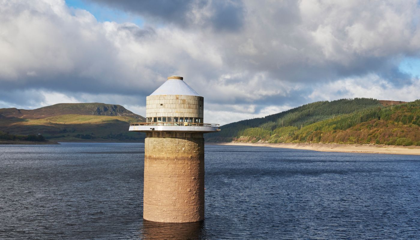 The water tower of Llyn Celyn reservoir stands in the foreground, with the undulating hills of the Arenig mountains stretching into the distance under a partly cloudy sky, showcasing the expansive beauty of Wales' largest reservoir.