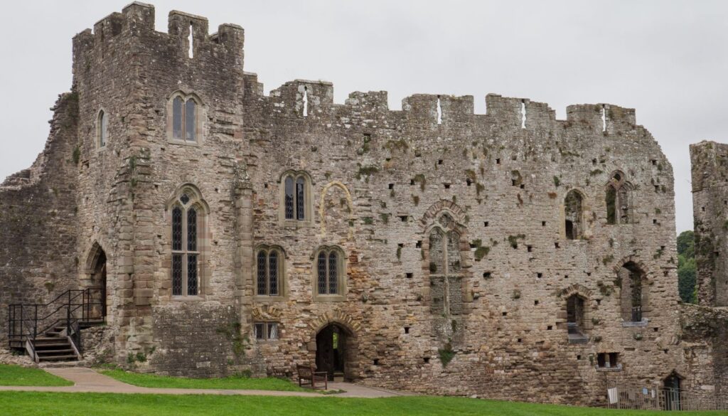 Ruins of Chepstow Castle with tall, weathered stone walls featuring Gothic windows, set against a soft sky, surrounded by a green lawn.