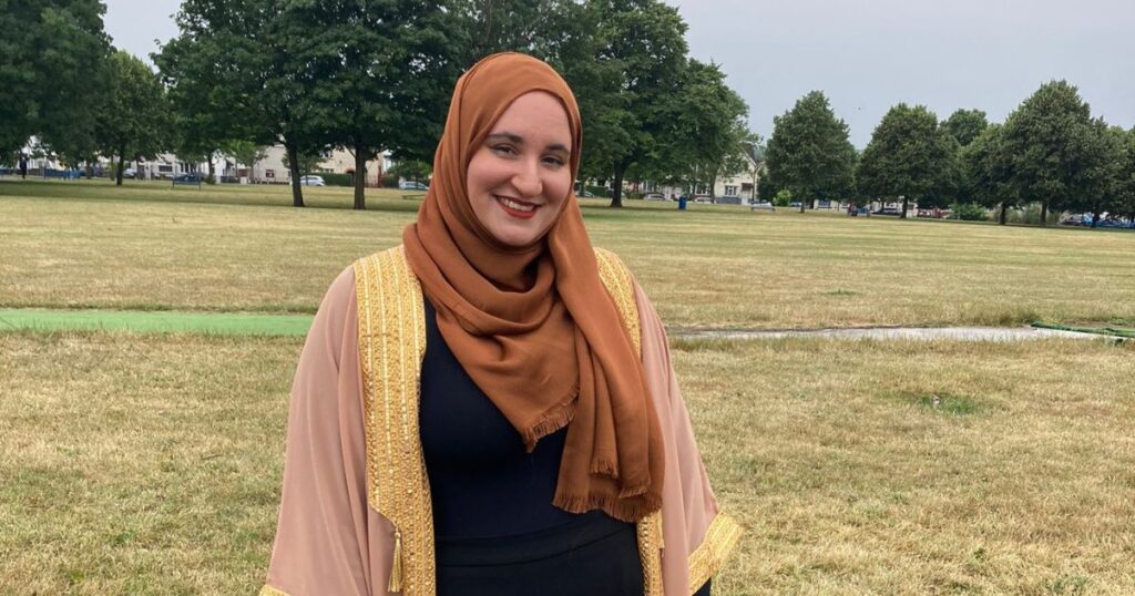 Hanan Issa, the National Poet of Wales, smiling warmly in a Cardiff park, adorned in a traditional brown hijab and a beige dress with golden trimmings, embodying the multicultural essence of modern Wales.