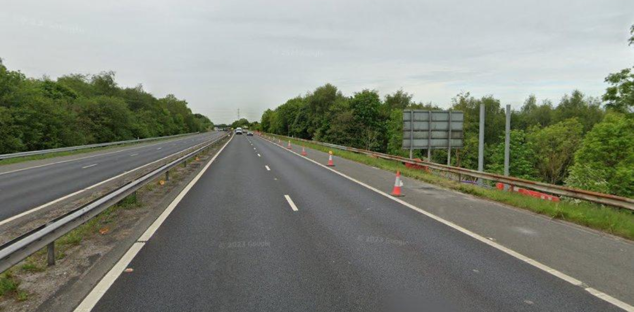 View of the M65 motorway showing a quiet section between Hapton and Burnley, with traffic cones hinting at lane closures or road work ahead, surrounded by lush greenery on a cloudy day.