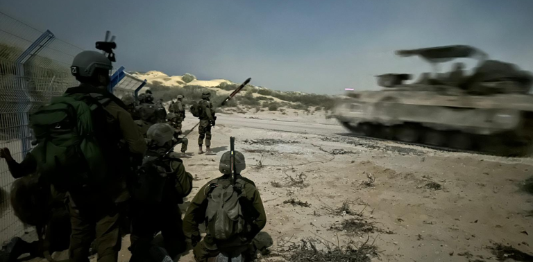 A group of soldiers in tactical gear and helmets observes as a tank speeds past them in a desert landscape, likely in Northern Israel, amidst a tense military scenario.