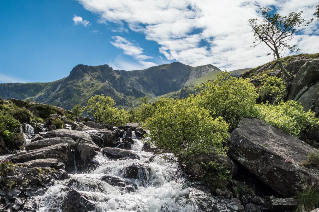 Waterfall flowing from Llyn Idwal at Cwm Idwal, surrounded by rocky slopes and green trees in Nant Ffrancon Valley, Snowdonia National Park.