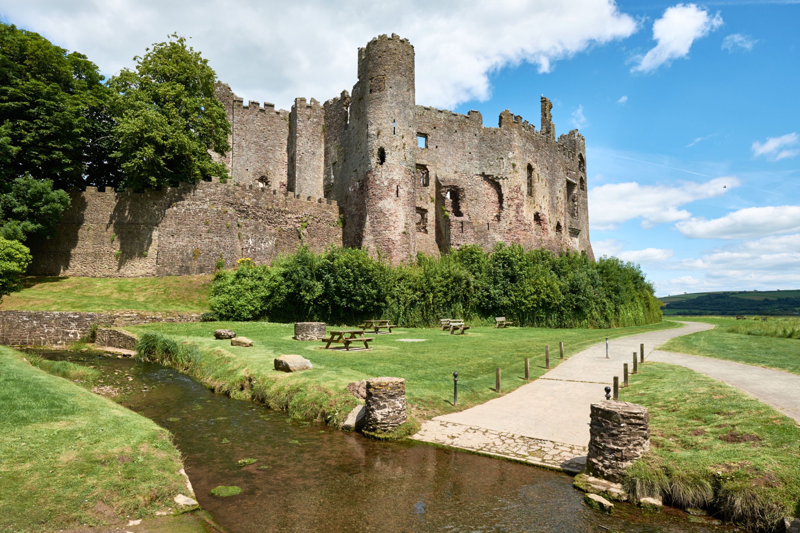 Ruins of Laugharne Castle in Carmarthenshire, Wales, with a river stream and picnic area in the foreground.