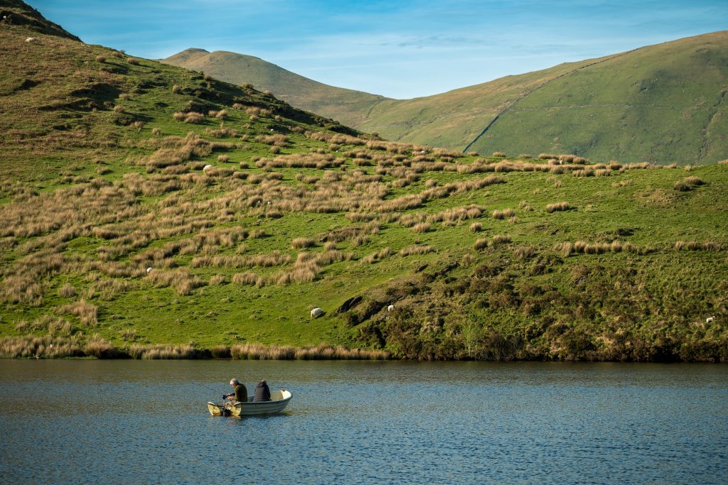 Two anglers fishing from a boat on Llyn Dywarchen in Eryri National Park, with the peak of Yr Wyddfa visible in the background.